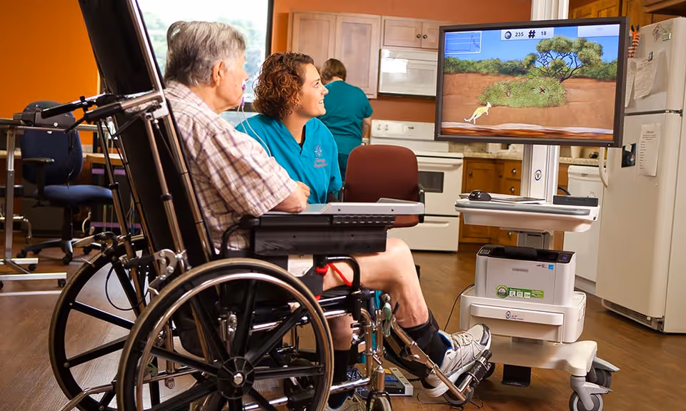 An elderly man in a wheelchair is playing a video game on a large screen with the assistance of a female caregiver in a teal uniform. The setting appears to be a rehabilitation or healthcare center with a kitchen area in the background.