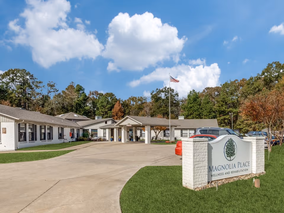 Entrance and front exterior of Magnolia Place Wellness and Rehabilitation with driveway, facility sign, flagpole, and parked cars.
