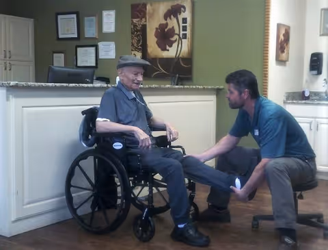 An elderly man sitting in a wheelchair smiling while a caregiver kneels in front of him, holding and examining his leg in a room with a reception desk and framed certificates on the wall.