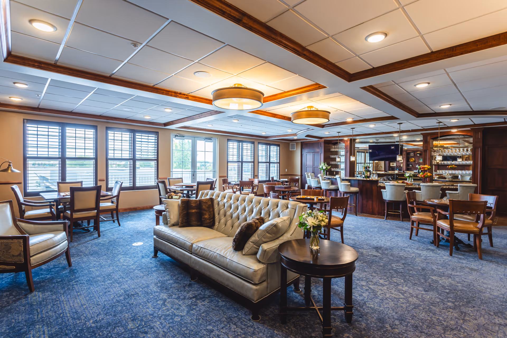 A spacious and well-lit common area with multiple seating options including a tufted leather sofa, armchairs, and wooden dining tables with chairs. The room features large windows with blinds, a carpeted floor, a ceiling with recessed lighting and wooden beams, and a bar area with stools and shelves stocked with glassware and bottles.