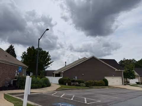 Parking area in front of single-story brick residential villas with shrubs and a cloudy sky overhead.
