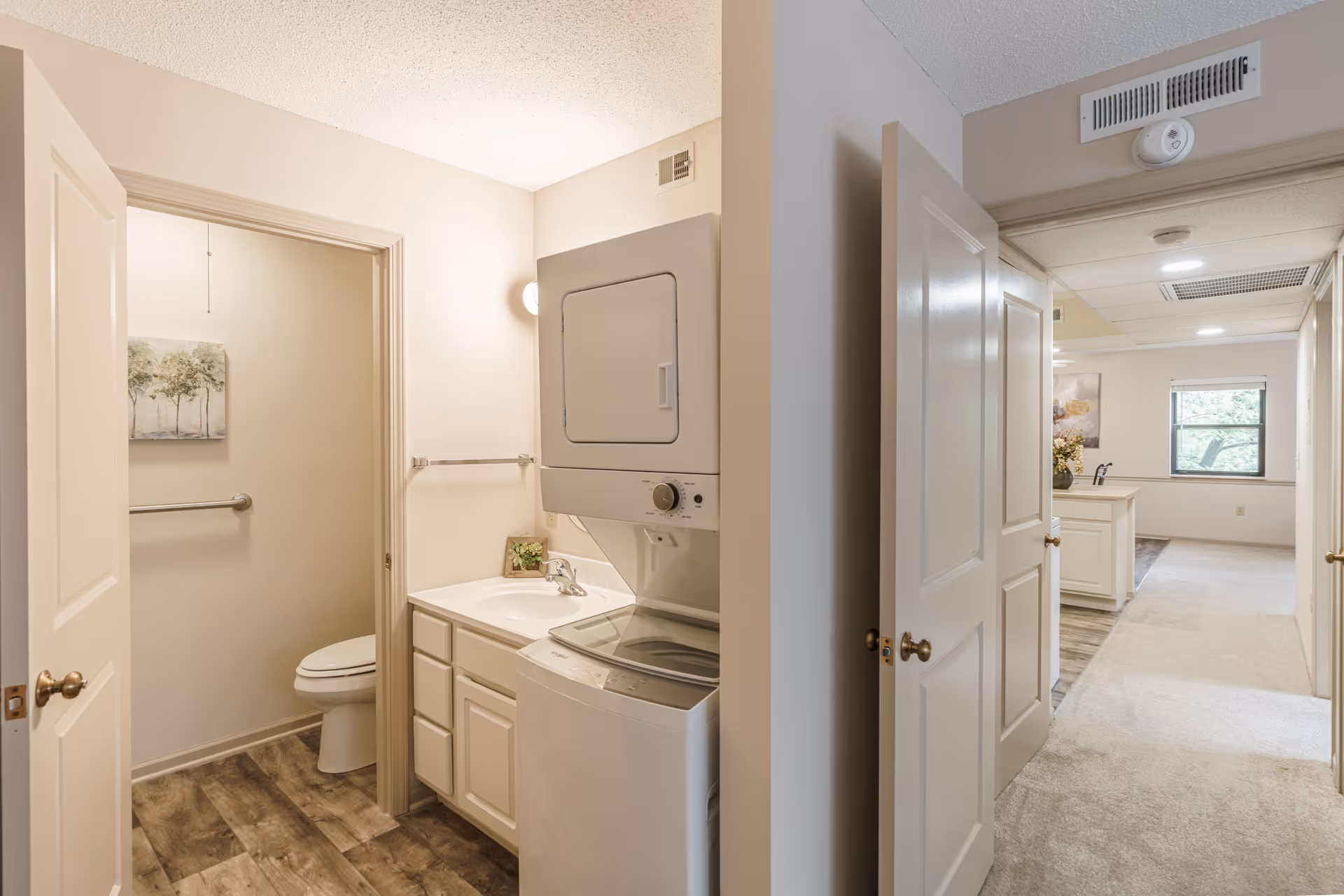 Interior view of a senior living facility showing a small laundry area with a stacked washer and dryer next to a sink and cabinet. To the left, there is a bathroom with a toilet, grab bar, and a painting on the wall. To the right, a hallway leads to a well-lit room with a window and carpeted floor.