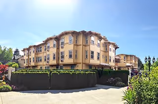 Exterior view of a multi-story senior living facility building with beige walls and multiple windows, surrounded by greenery and a clear blue sky.
