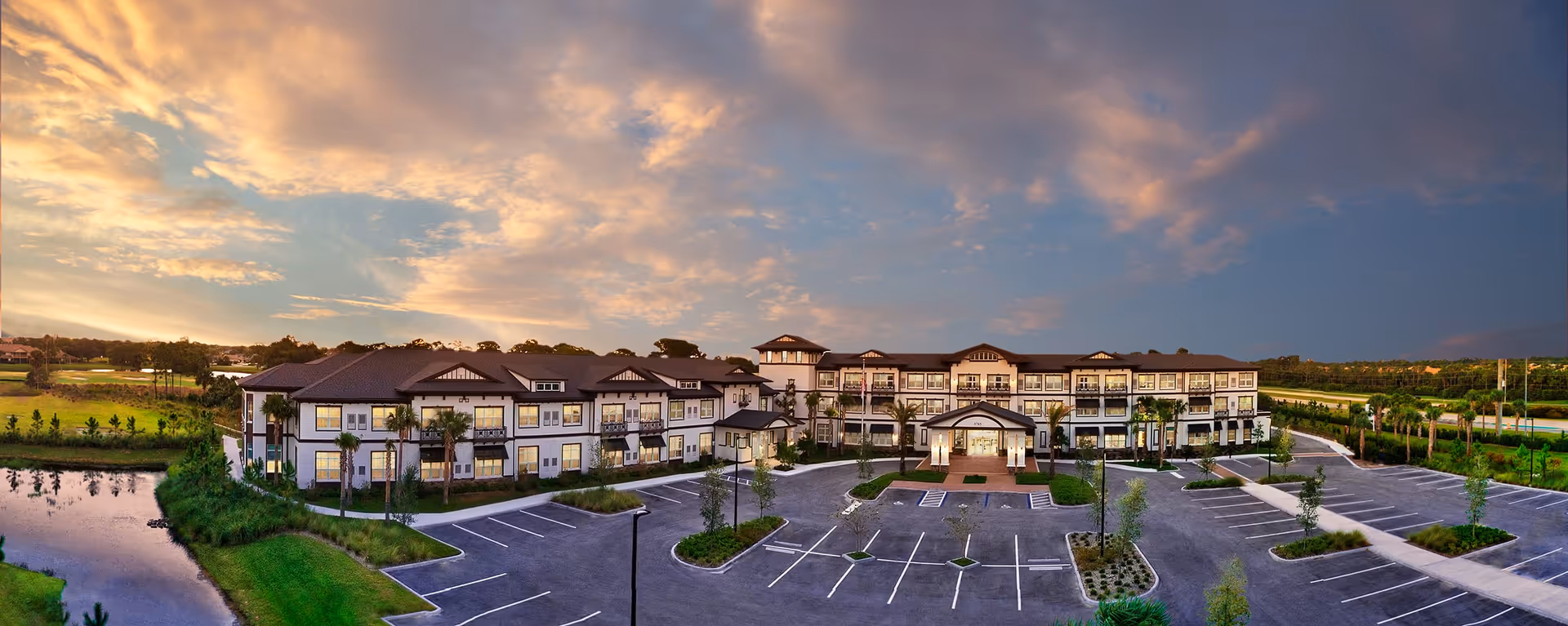 Panoramic view of Discovery Commons Hobe Sound, a large senior living facility building with multiple balconies and a covered entrance, surrounded by landscaped greenery and an empty parking lot under a colorful sunset sky.
