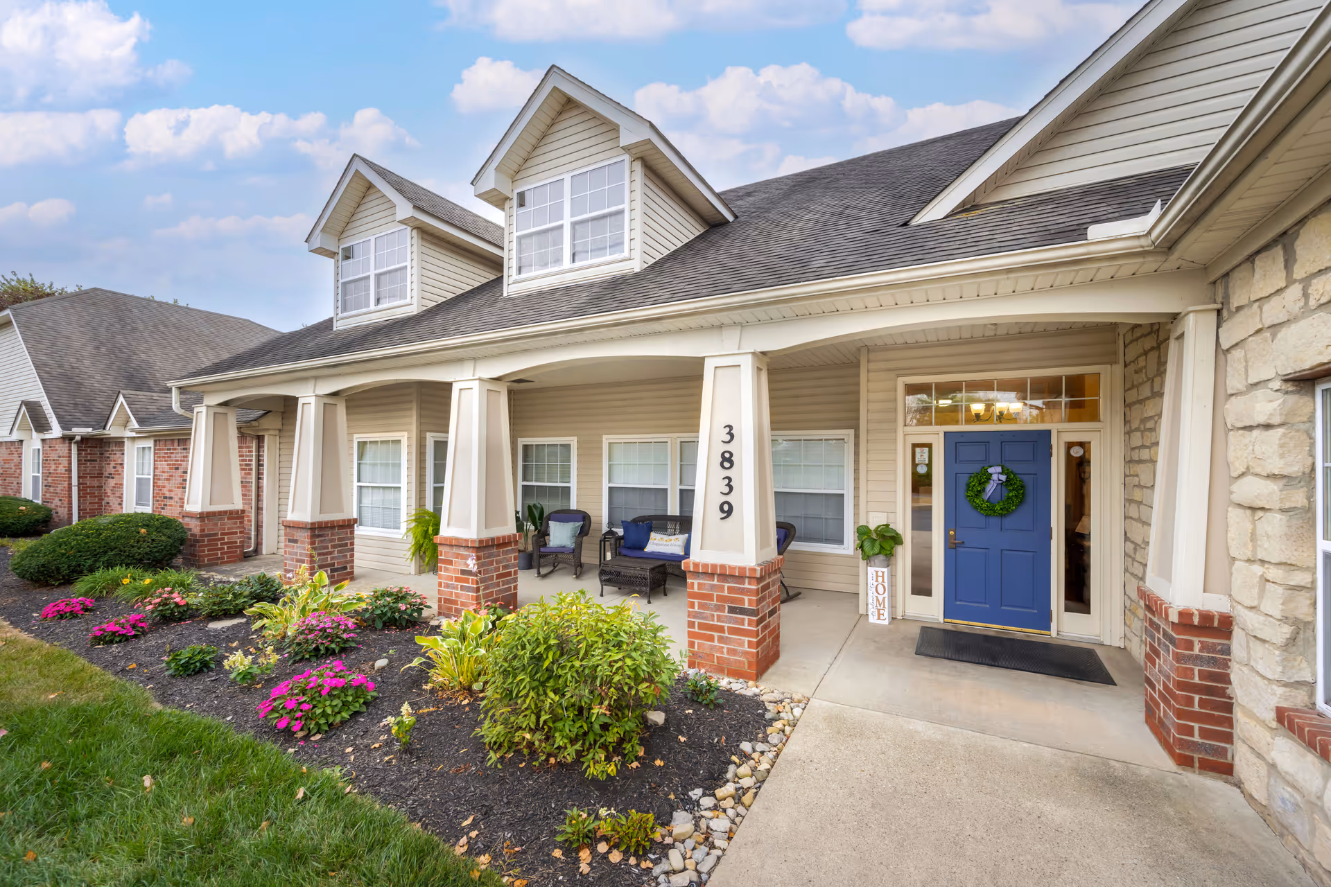 Front exterior view of a senior living facility with a covered porch, blue front door decorated with a green wreath, brick and beige siding, and landscaped flower beds with green shrubs and pink flowers.