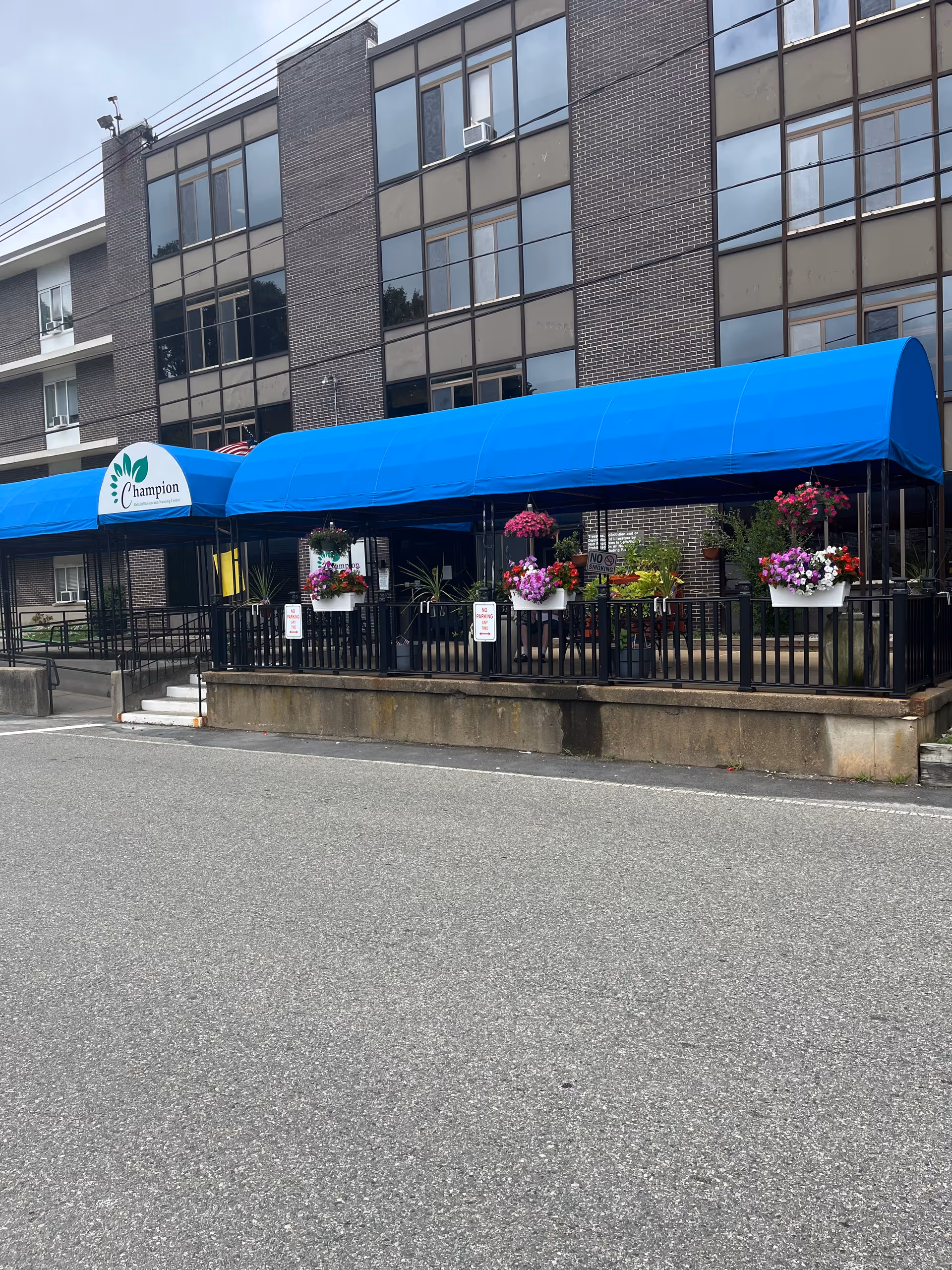 Exterior view of Champion Rehabilitation and Nursing Center showing a multi-story brick building with large windows. A blue awning covers the entrance area, which is decorated with hanging flower pots containing colorful flowers. There is a paved area in front of the building.