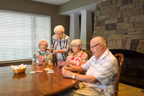 Four elderly individuals sitting around a wooden table playing cards in a cozy room with a stone fireplace and large window with blinds.