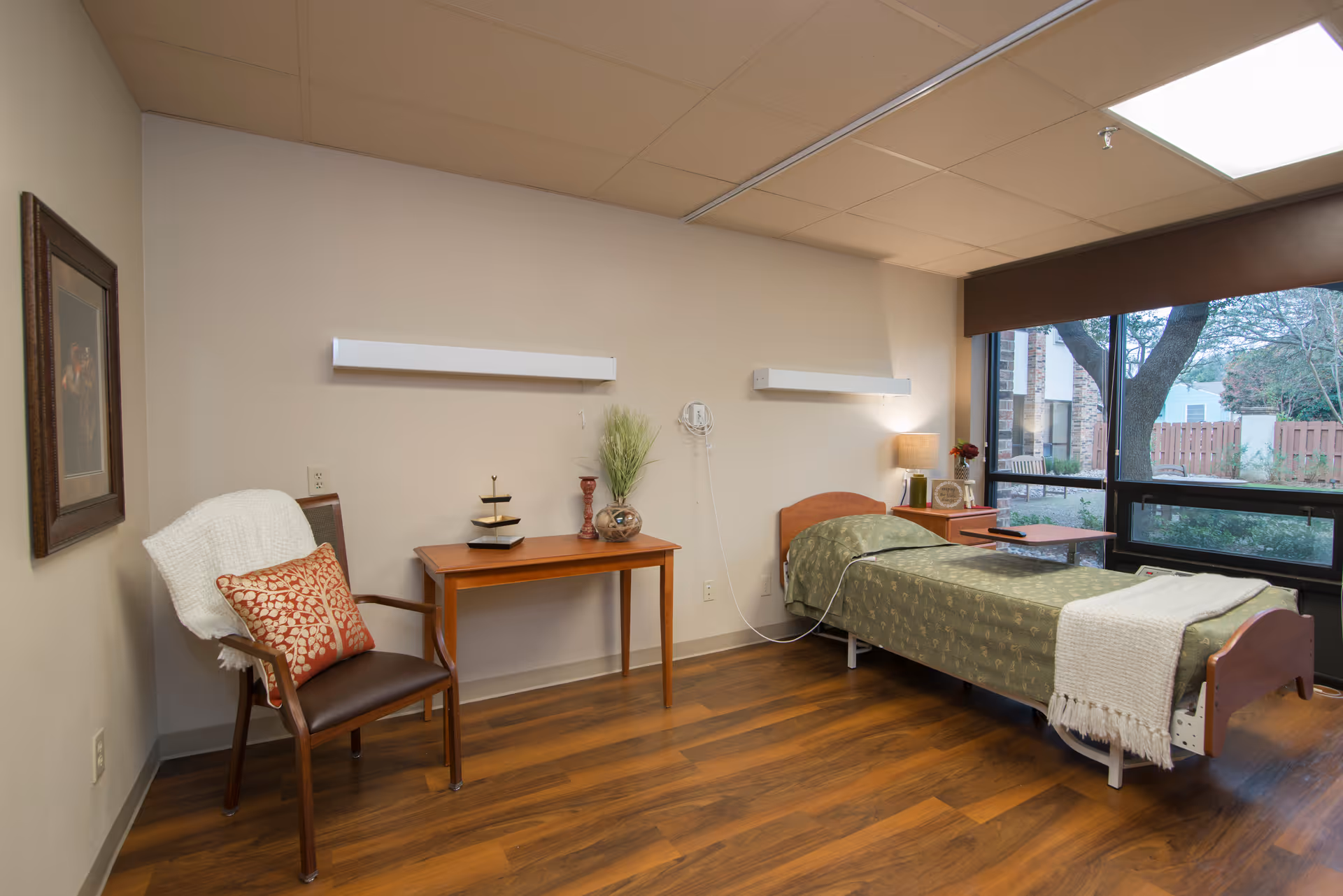 A cozy senior living bedroom with a single bed covered in a green patterned blanket and a white throw. Next to the bed is a wooden nightstand with a lamp, clock, and a small vase of flowers. A wooden table with decorative items and a chair with a white throw and red patterned pillow are also in the room. Large windows provide a view of trees and a fenced yard outside. The room has wooden flooring and beige walls.