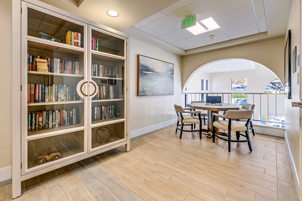 Bright senior living common area with a glass-front bookcase and a round table with chairs by an arched railing.