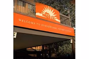 Orange sign with a white sun and building logo above the text 'WELCOME TO STONEBRIDGE TOWN CENTER' mounted on a wooden structure with trees in the background.