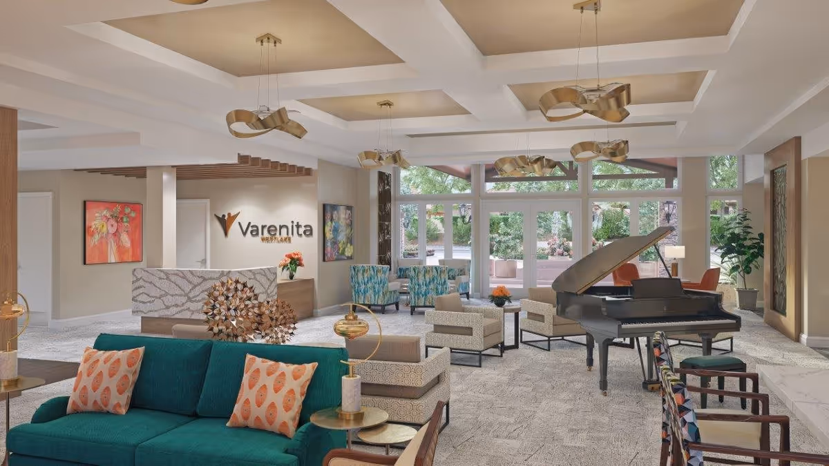 A spacious and well-lit living room area in Varenita Westlake featuring modern furniture including a teal sofa with patterned pillows, beige armchairs, a grand piano, and large windows overlooking a garden. The ceiling has decorative beams and unique pendant lights, with artwork on the walls and plants adding to the decor.