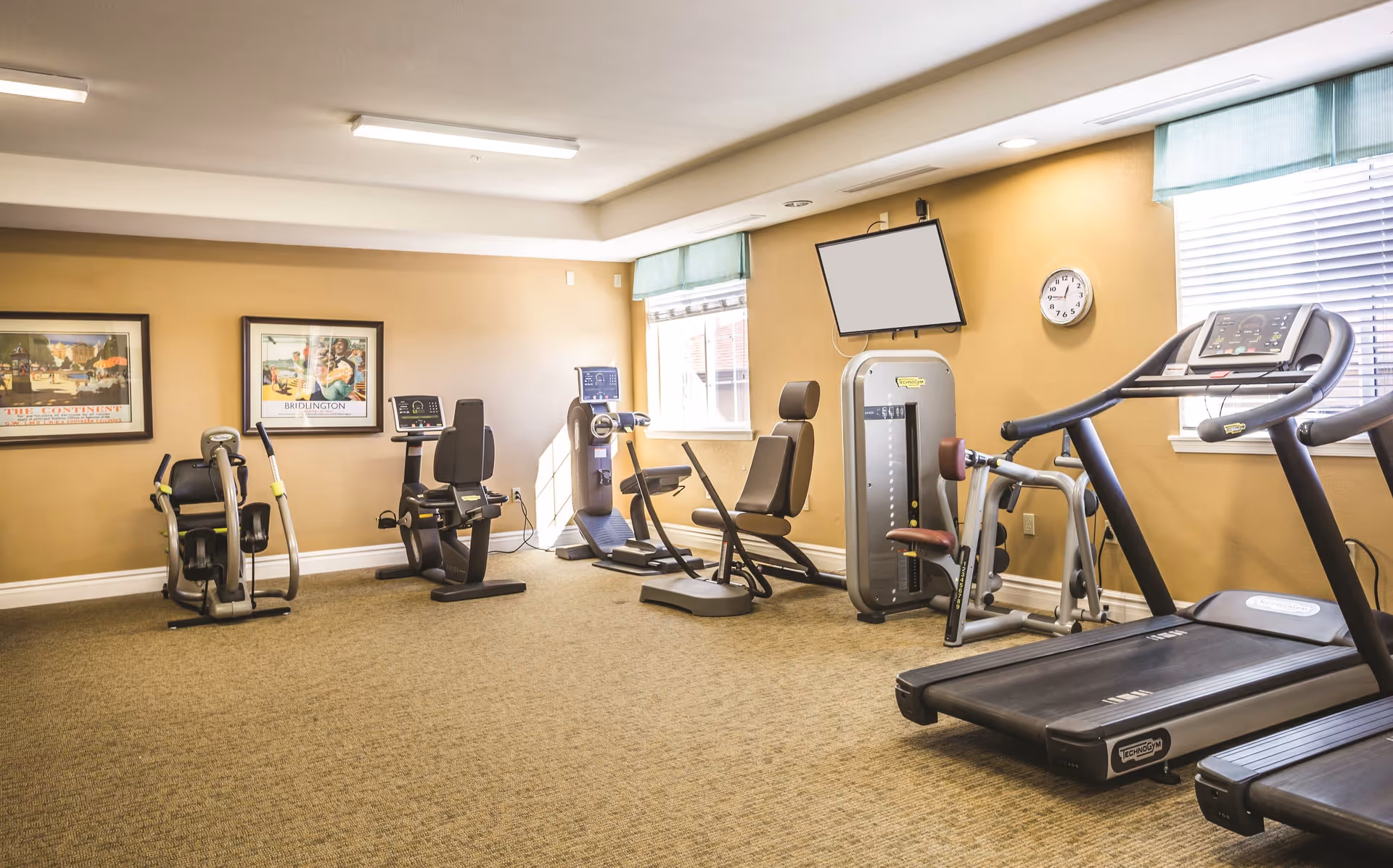 A well-lit exercise room with various fitness equipment including a treadmill, stationary bikes, and strength training machines. The room has beige walls, carpeted floor, two windows with blinds, two framed pictures on the wall, a wall clock, and a mounted TV screen.