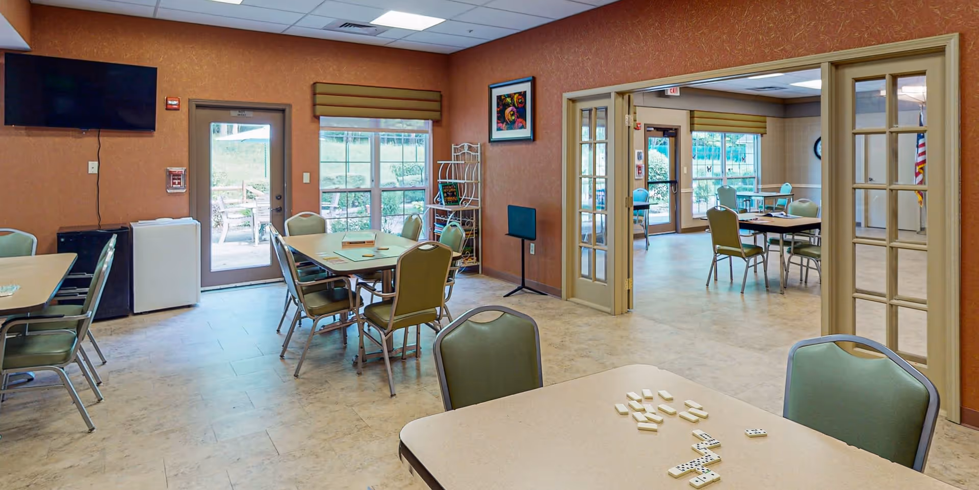 Interior view of a community room with multiple tables and chairs arranged for group activities. One table in the foreground has dominoes laid out. The room has large windows and glass doors letting in natural light, with a TV mounted on the wall and a small refrigerator in the corner. Another adjoining room with similar setup is visible through glass-paneled double doors.