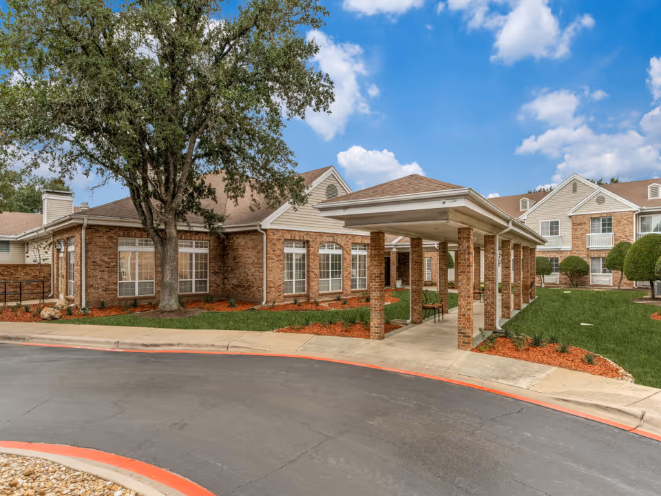 Exterior view of Juniper Village at Spicewood Summit showing a brick building with large windows, a covered walkway supported by brick columns, neatly trimmed bushes, a large tree, and a curved driveway with a red curb under a partly cloudy blue sky.