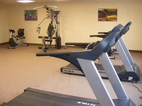 Small fitness room with treadmills in the foreground, a recumbent bike and a multi-station weight machine against a beige wall with framed artwork.