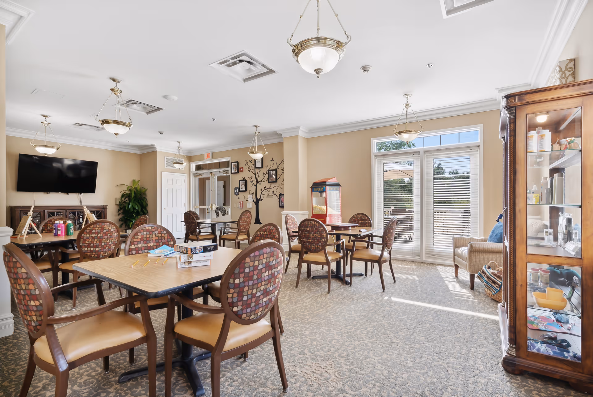 A bright and spacious common area in Bear Creek Assisted Living featuring multiple tables and chairs with patterned upholstery, a large flat-screen TV mounted on the wall, a popcorn machine near the windows, and a glass display cabinet with various items. The room has beige walls, carpeted floors, and large windows letting in natural light.