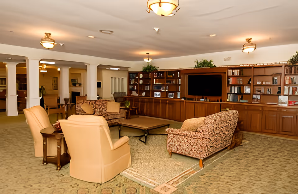 A spacious living room area with beige carpet and multiple upholstered chairs and sofas arranged around a central coffee table. The back wall features built-in wooden shelves filled with books and decorative items, and a large flat-screen TV is mounted in the center. The room is softly lit by ceiling lights and has white columns dividing the space.