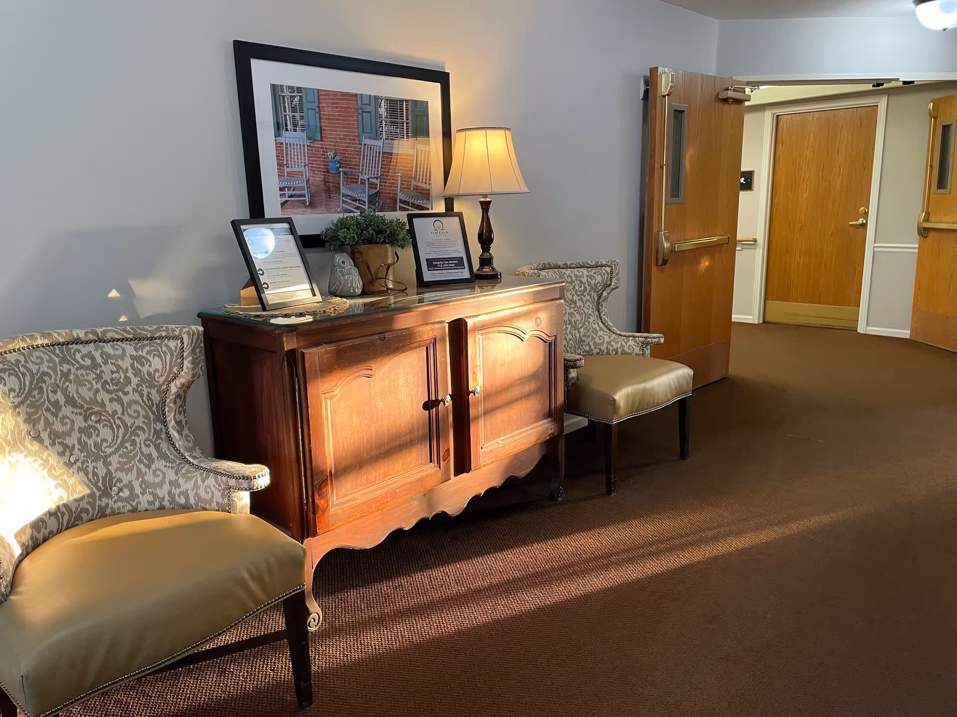 A hallway area in a senior living facility with two upholstered chairs flanking a wooden cabinet. On top of the cabinet are two framed documents, a small potted plant, a decorative owl, and a table lamp. Above the cabinet hangs a framed picture of rocking chairs on a porch. The hallway has brown carpet and wooden doors, with one door open showing another hallway.