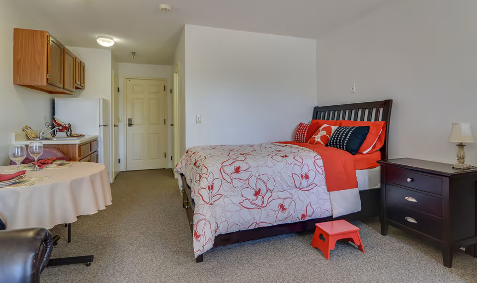 A studio apartment in a senior living facility featuring a bed with red and white floral bedding and multiple pillows, a dark wooden nightstand with a lamp, a small red step stool, a round dining table set with two place settings and wine glasses, a kitchenette with wooden cabinets, a refrigerator, and a countertop with a decorative tricycle, and a closed door at the end of the room.