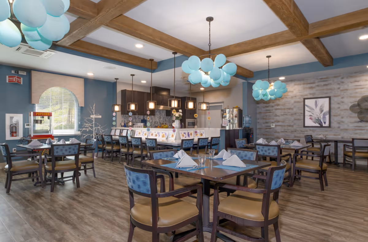 A spacious dining room in a senior living facility with multiple tables and chairs arranged neatly. The tables are set with folded napkins and glassware. The room features wooden beams on the ceiling, modern pendant lights, and decorative blue balloon-like light fixtures. There is a popcorn machine near a window with an arched valance, and a kitchen area with a counter and bar stools in the background. The walls have a mix of blue paint and textured wallpaper with framed artwork.