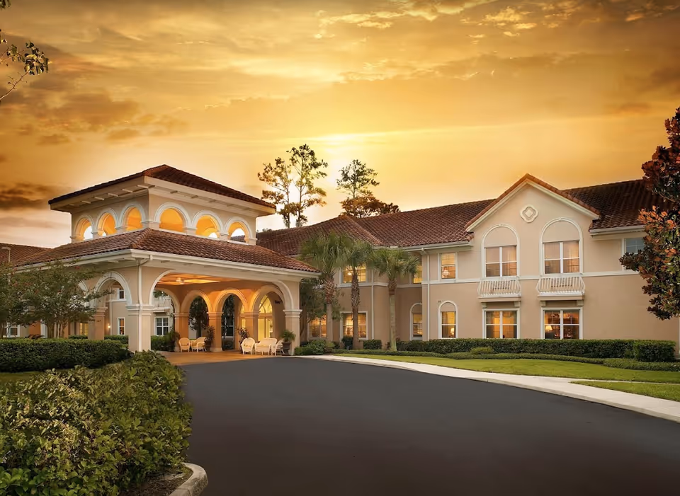Exterior view of The Cove at Marsh Landing senior living facility at sunset, featuring a large beige building with arched windows and a covered entrance with arches, surrounded by manicured bushes, palm trees, and a curved driveway.