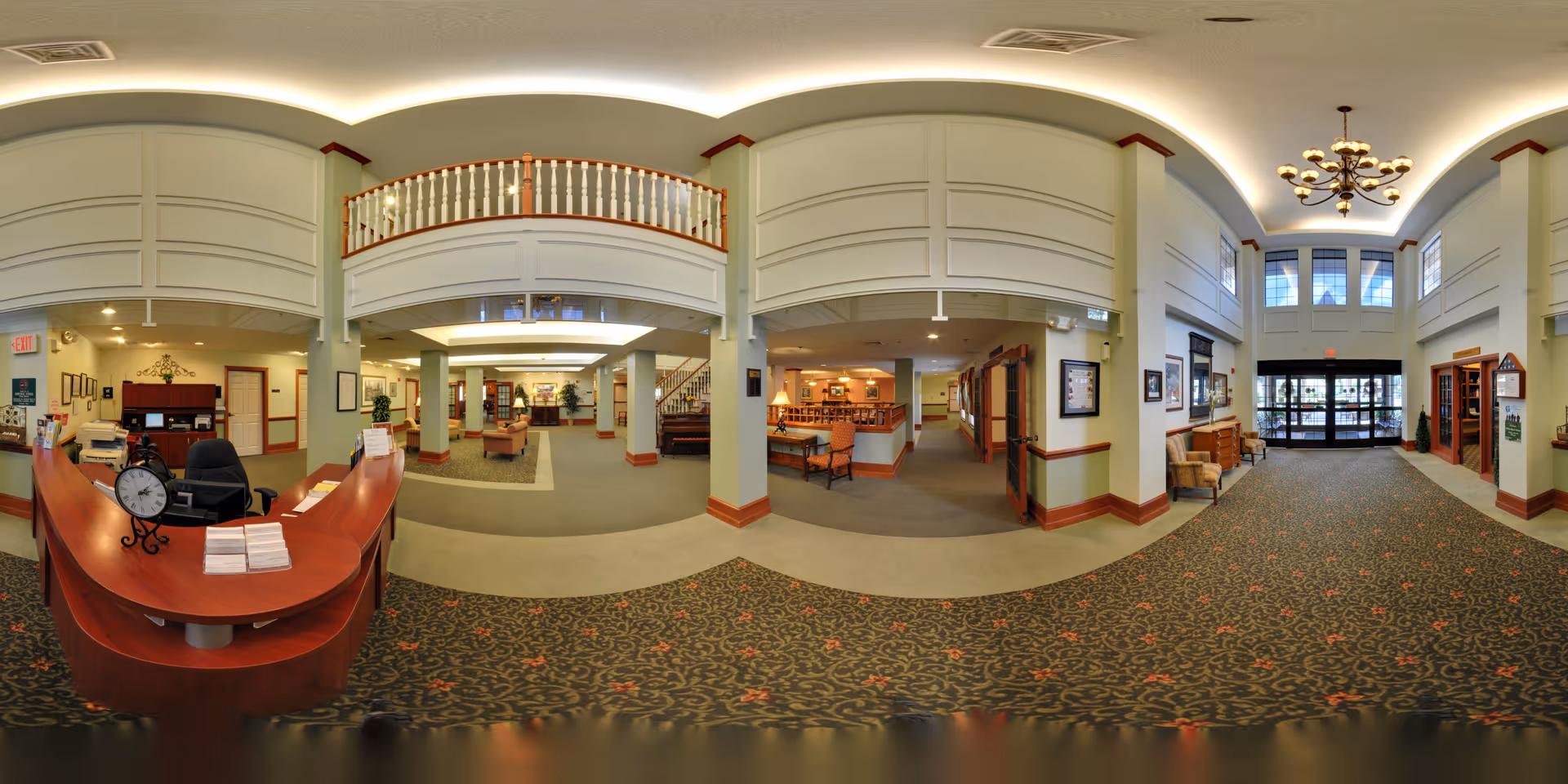 Wide panoramic view of a senior living facility lobby area featuring a curved wooden reception desk with a clock and brochures, seating areas with armchairs and tables, decorative plants, and a high ceiling with a chandelier. The entrance doors are visible at the far end, with large windows above allowing natural light to fill the space. The interior design includes carpeted floors with a floral pattern, light-colored walls with wood trim, and a balcony railing on the upper level.