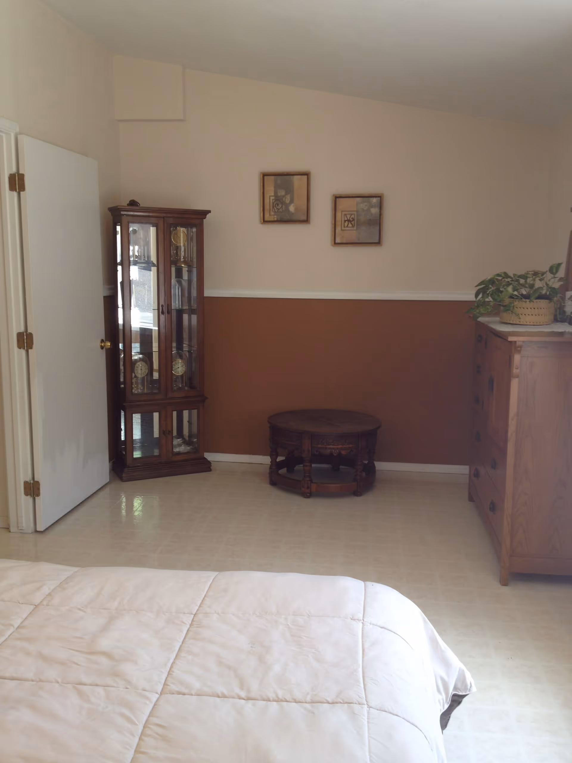 A bedroom with a beige and brown color scheme. The foreground shows a bed with a light beige comforter. Against the far wall, there is a wooden display cabinet with glass doors containing clocks, a round wooden coffee table, and a wooden dresser with a potted plant on top. Two framed abstract artworks hang on the wall above the coffee table. The floor is covered with light-colored tiles, and a white door is partially open on the left side.