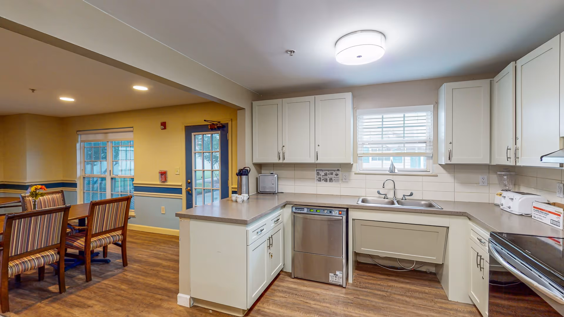 A clean and well-lit kitchen area with white cabinets, a double sink under a window with blinds, a dishwasher, and various small appliances on the countertops. Adjacent to the kitchen is a dining area with a wooden table and striped upholstered chairs, with windows and a door leading outside.
