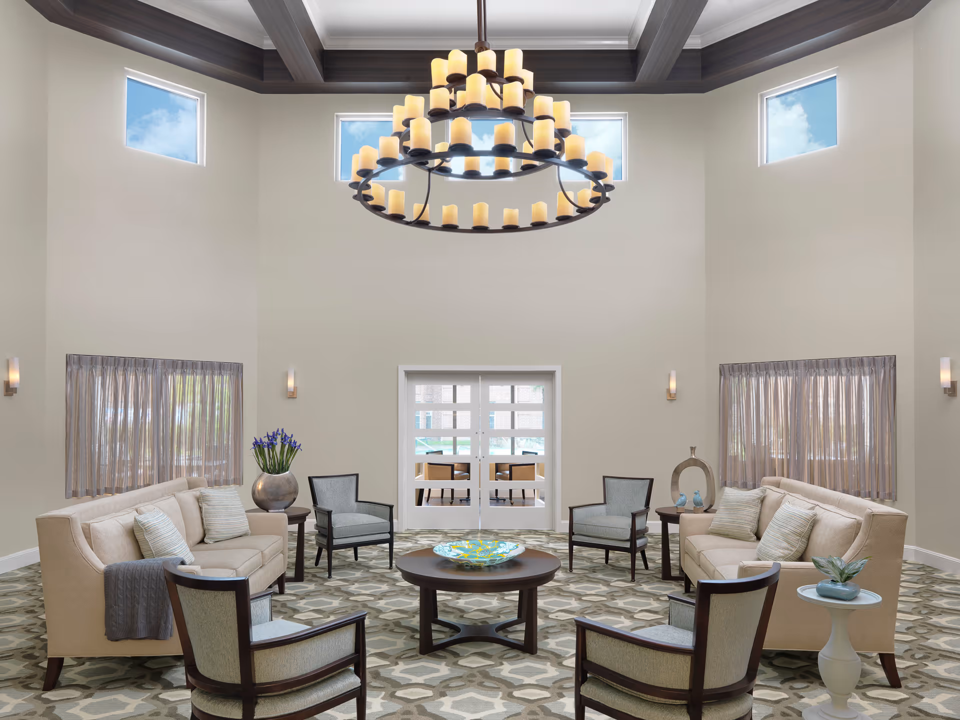 A spacious and well-lit living room with high ceilings and a large circular chandelier with multiple candle-like lights. The room features two beige sofas facing each other, four armchairs, and three round wooden tables, one with a decorative glass bowl and others with plants and ornaments. There are four small windows near the ceiling and two larger windows with sheer curtains on either side of a double glass door leading to another room with additional seating.