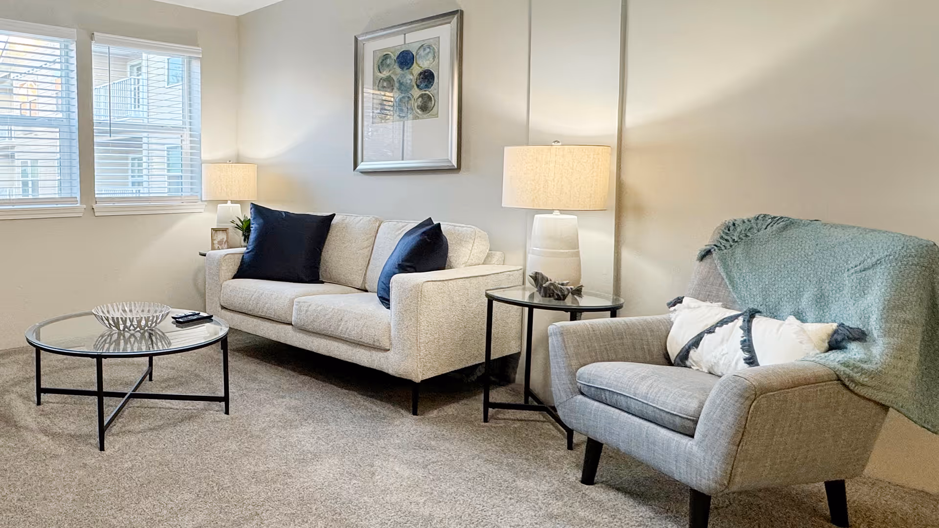 Bright living room with a sofa, armchair draped with a throw, glass coffee table, side tables with lamps, and framed wall art beneath windows.
