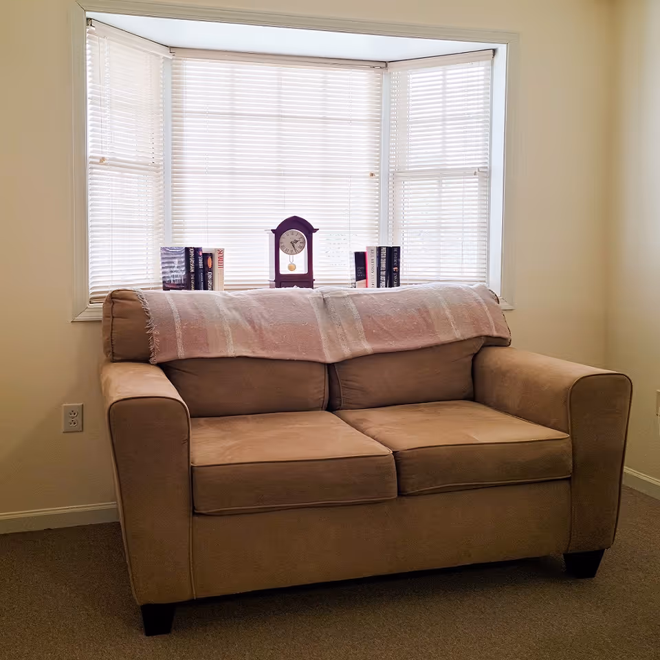 A beige loveseat sofa with a light pink throw blanket draped over the backrest, positioned in front of a bay window with closed white blinds. On the window sill behind the sofa, there is a small purple clock and several books arranged on either side of the clock. The room has light-colored walls and carpeted flooring.