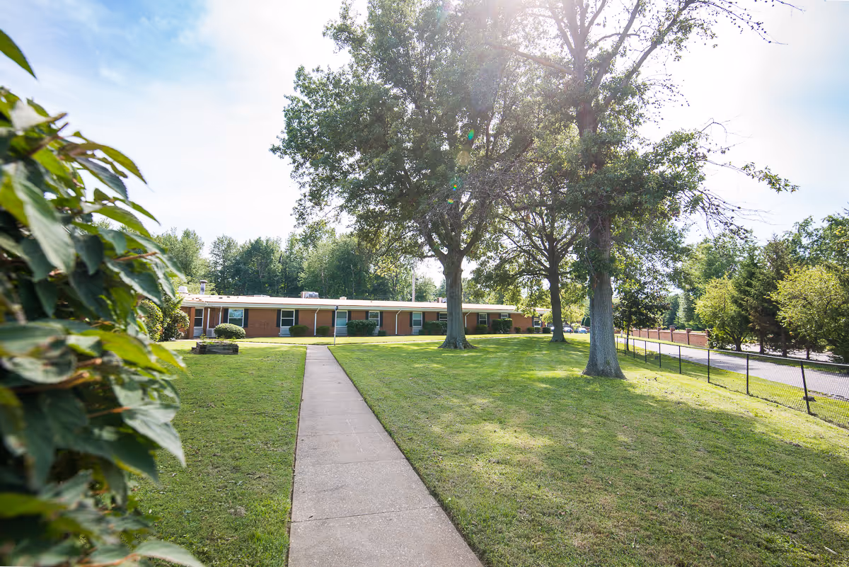 A single-story brick building with multiple windows and doors, surrounded by a well-maintained lawn with several large trees. A concrete walkway leads from the foreground to the building entrance. The sky is partly cloudy with sunlight filtering through the trees.