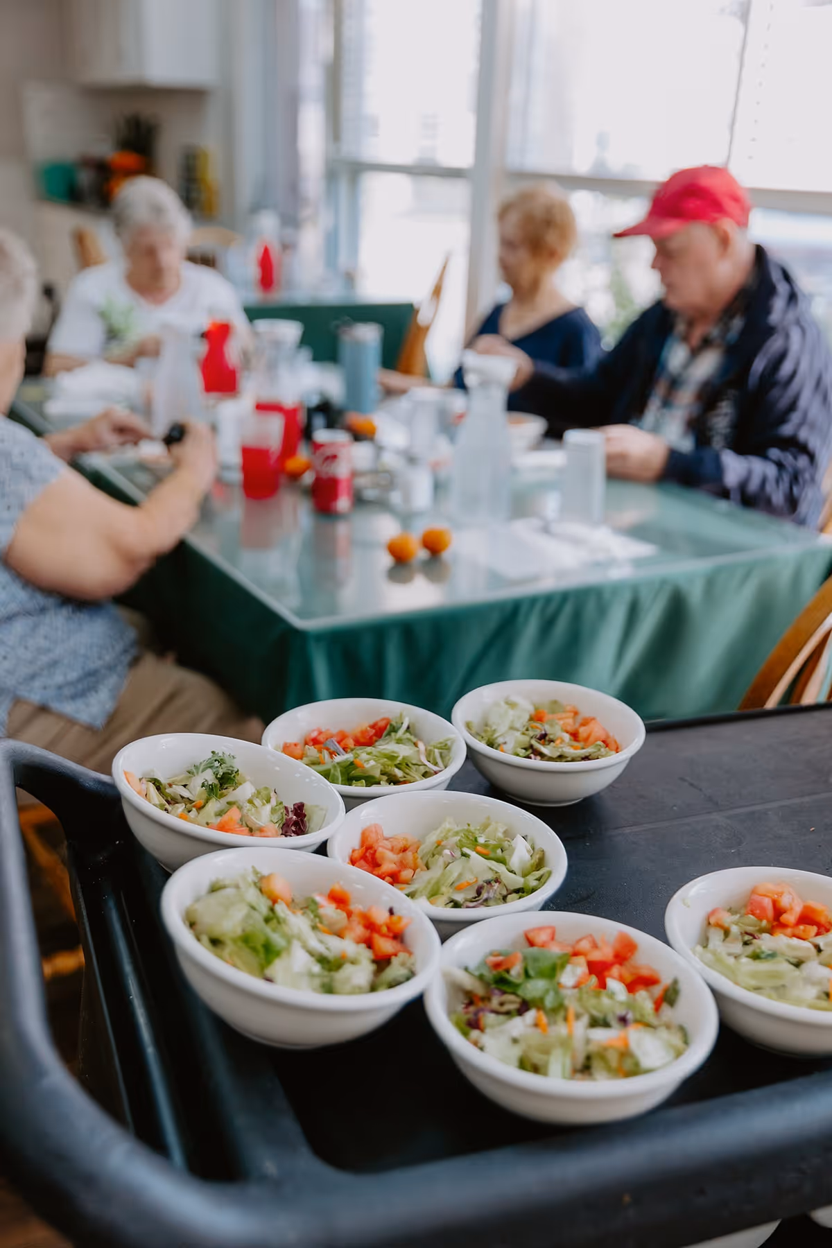 Several bowls of fresh salad with lettuce, tomatoes, and other vegetables on a serving cart in the foreground. In the background, a group of elderly people are seated around a table covered with a green tablecloth, engaging in conversation and dining in a bright room with large windows.