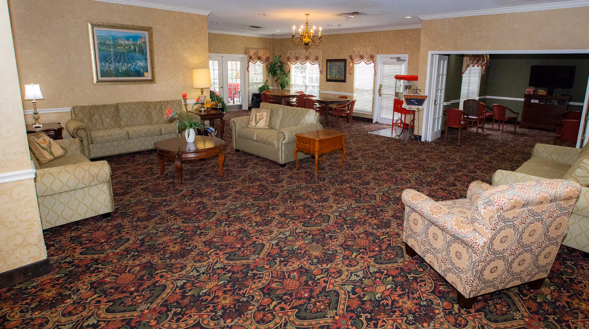 A spacious living room area in an assisted living facility featuring patterned carpet, multiple upholstered sofas and armchairs, wooden coffee and side tables, a chandelier, framed artwork on the walls, and large windows with curtains letting in natural light. In the background, there is a dining area with tables and chairs, and a popcorn machine near a doorway leading to another room with a television and additional seating.