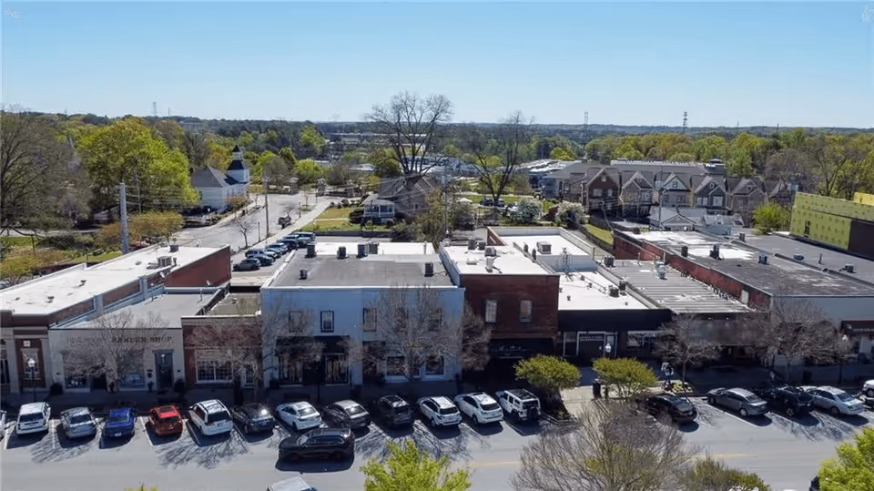 Aerial view of a small town commercial area with a row of buildings and parked cars along the street. Trees and residential houses are visible in the background under a clear blue sky.