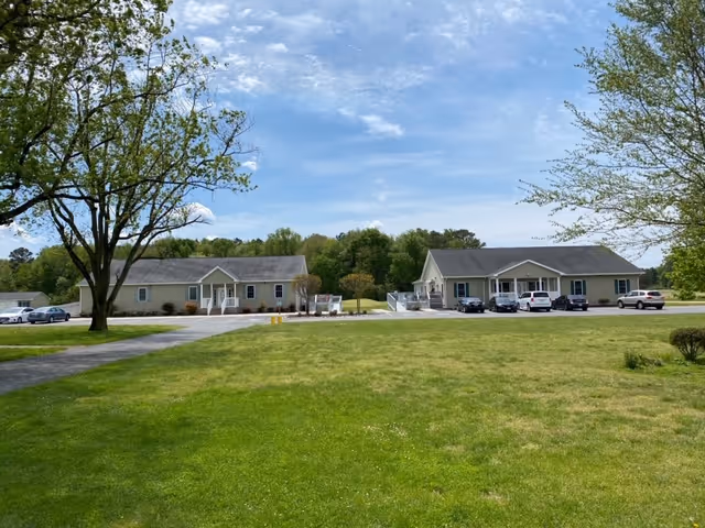 Two single-story beige buildings with gray roofs situated on a large grassy lawn with a few trees and several parked cars in front. The sky is partly cloudy with blue patches visible.