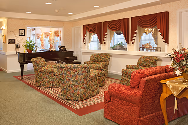 A cozy living room area with floral patterned armchairs and a red upholstered armchair arranged around a rug. There is a grand piano in the background near a window with red valances and white blinds. A wooden side table with a floral arrangement is visible on the right side.