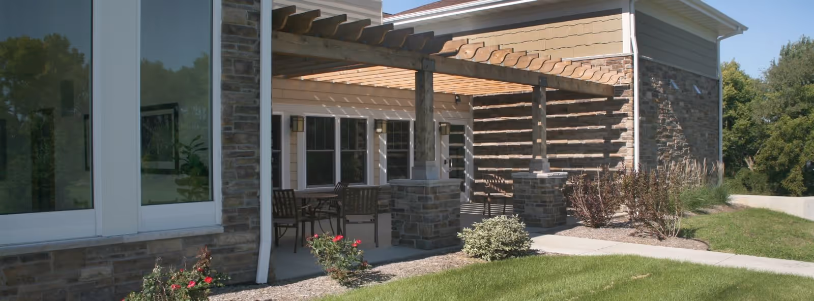 Outdoor patio area at Oak Park Place Janesville featuring a wooden pergola with stone pillars, outdoor seating with tables and chairs, surrounded by landscaped grass and plants.