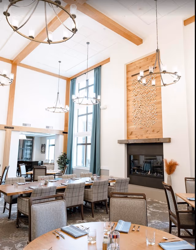 A bright and spacious dining room with high ceilings, wooden beams, and large windows with teal curtains. The room features multiple wooden tables set with napkins, glasses, and silverware, surrounded by cushioned chairs. There are modern circular chandeliers hanging from the ceiling and a fireplace with a decorative wooden panel above it on one wall.