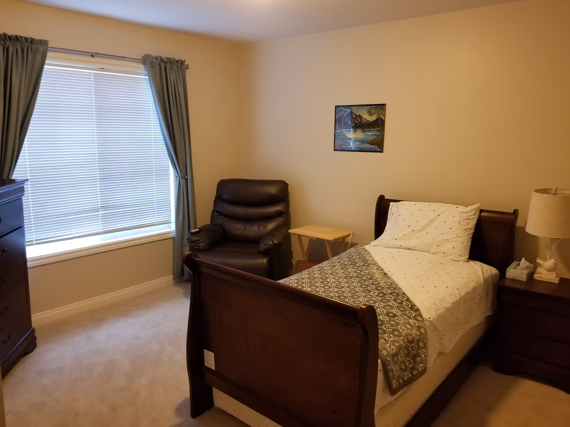 A cozy bedroom with a single wooden bed made with white and gray bedding. Next to the bed is a wooden nightstand with a lamp, tissue box, and decorative item. There is a dark brown leather recliner chair beside a small wooden folding table. A window with closed blinds and blue curtains is on the left wall, and a dresser is partially visible. A framed landscape painting hangs on the beige wall above the chair.