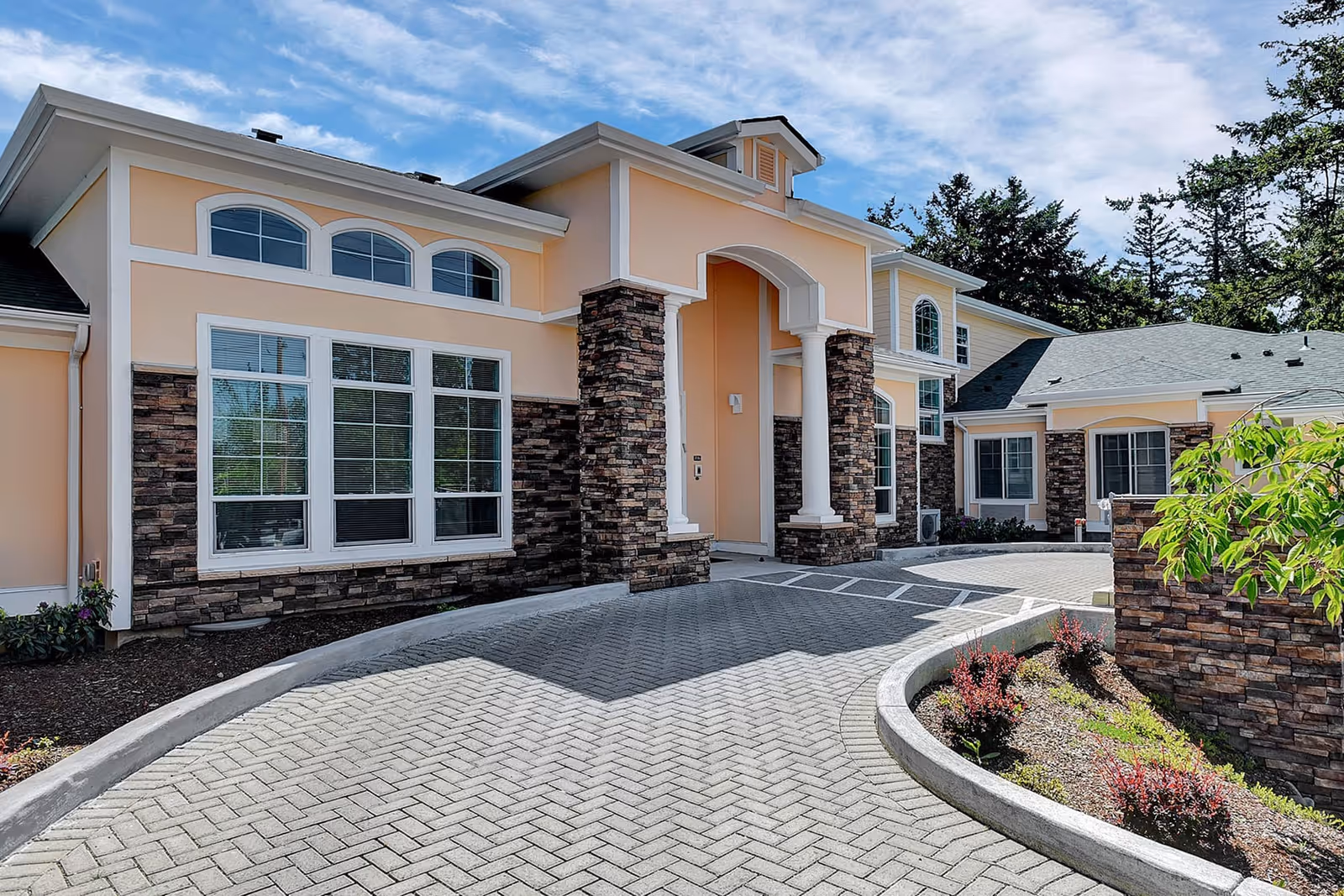 Exterior view of a senior living facility building with peach-colored walls, stone accents, large windows, and a covered entrance with white columns. The driveway is paved with interlocking bricks and curves around a landscaped area with small shrubs and plants. Trees and a partly cloudy sky are visible in the background.