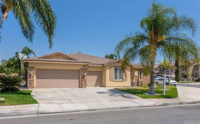 Single-story residential building with a three-car garage, beige exterior walls, and a tiled roof. The driveway and sidewalk are concrete, and there are palm trees and green lawns in front of the house. Other houses and parked cars are visible in the background under a clear blue sky.