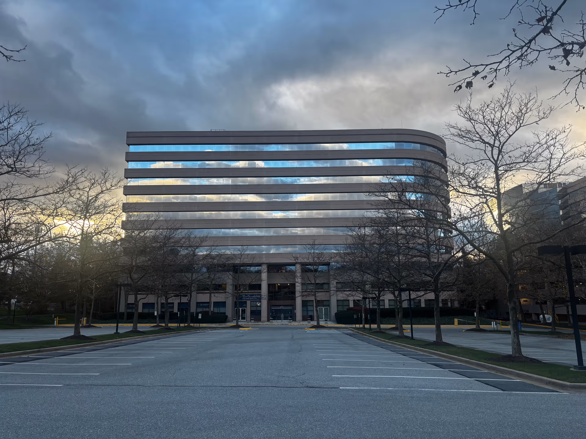 A modern multi-story office building with reflective glass windows showing the cloudy sky. The building is surrounded by leafless trees and an empty parking lot in the foreground during dusk or early evening.