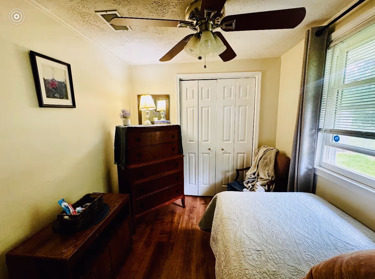 A small bedroom with a bed covered in a white quilt, a wooden dresser with a lamp and a small plant on top, a chair with a blanket draped over it, a window with blinds and gray curtains, and a ceiling fan with lights.