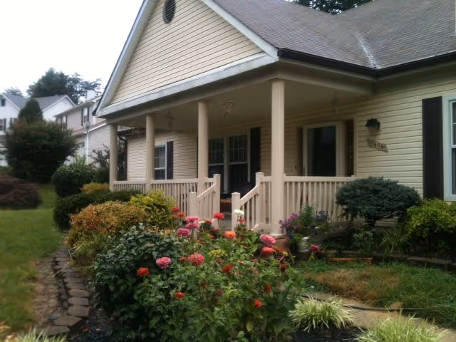 Front exterior view of a beige single-story house with a covered porch, surrounded by a garden with various green shrubs and colorful flowers.