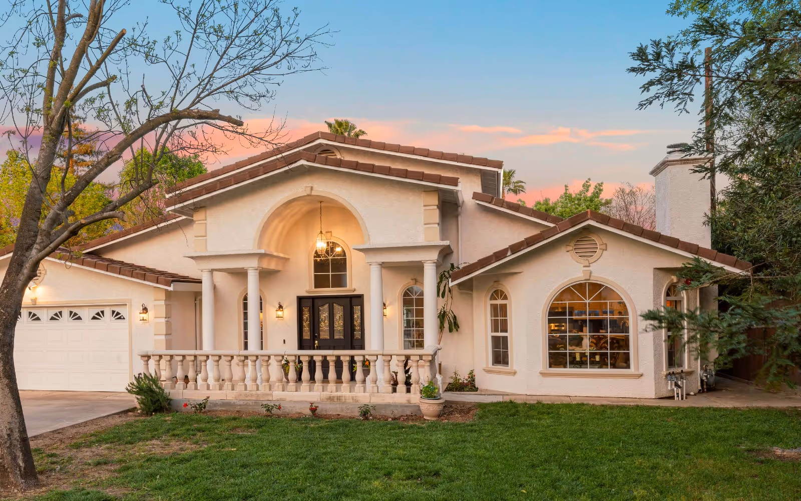 Front exterior of a Mediterranean-style two-story house with columns, arched windows, a garage, and a grassy lawn at sunset.