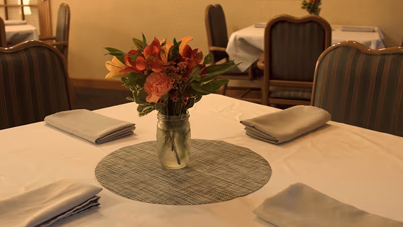 A dining table set with a white tablecloth, four folded napkins, and a centerpiece of colorful flowers in a glass jar. In the background, there are more tables and chairs with similar settings in a softly lit room.