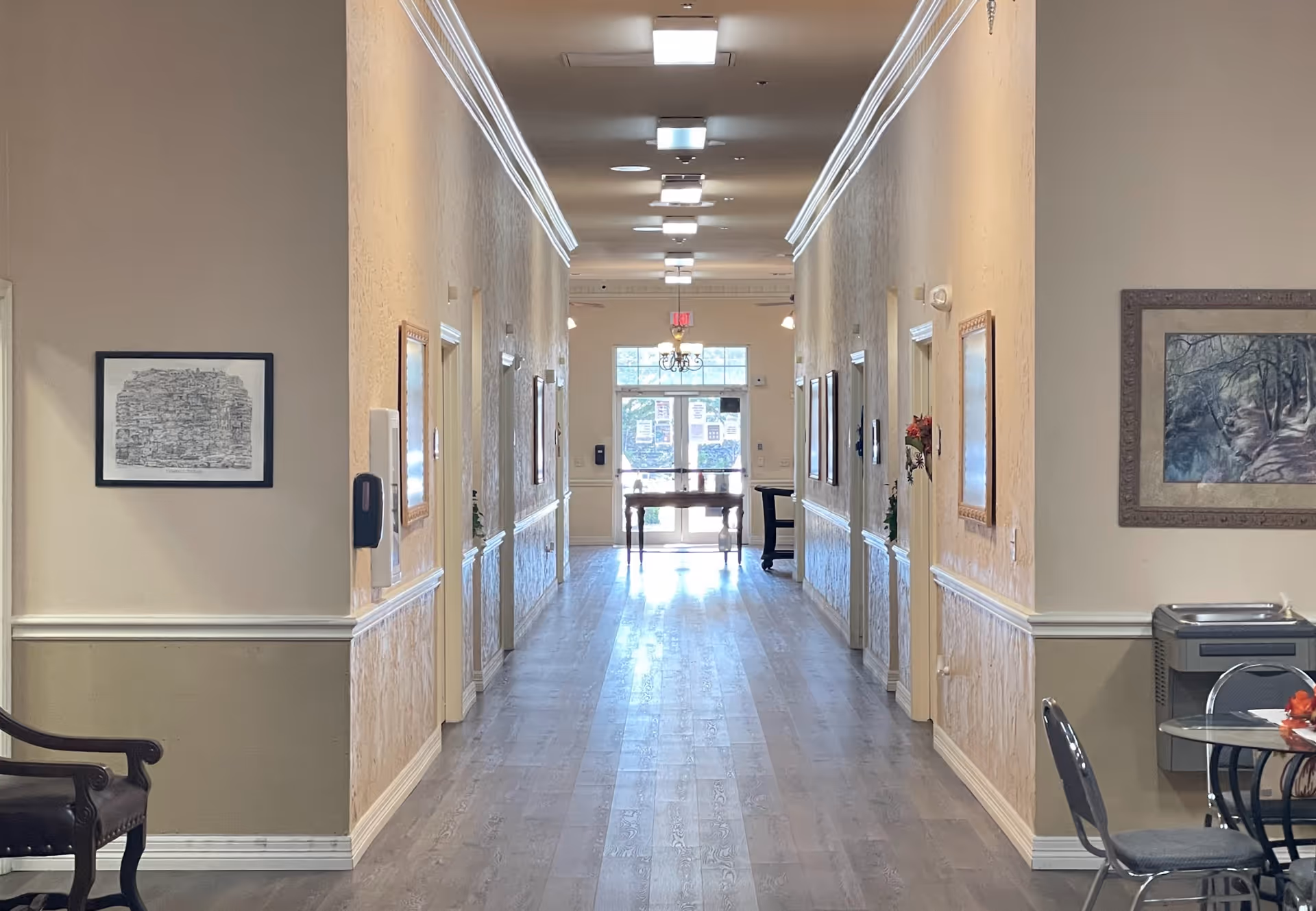 A well-lit hallway in an assisted living facility with light-colored walls and wood flooring. The hallway is decorated with framed pictures on the walls and has several doors along both sides. At the far end, there is a glass door letting in natural light. On the right side near the foreground, there is a small round table with chairs and a water fountain nearby.