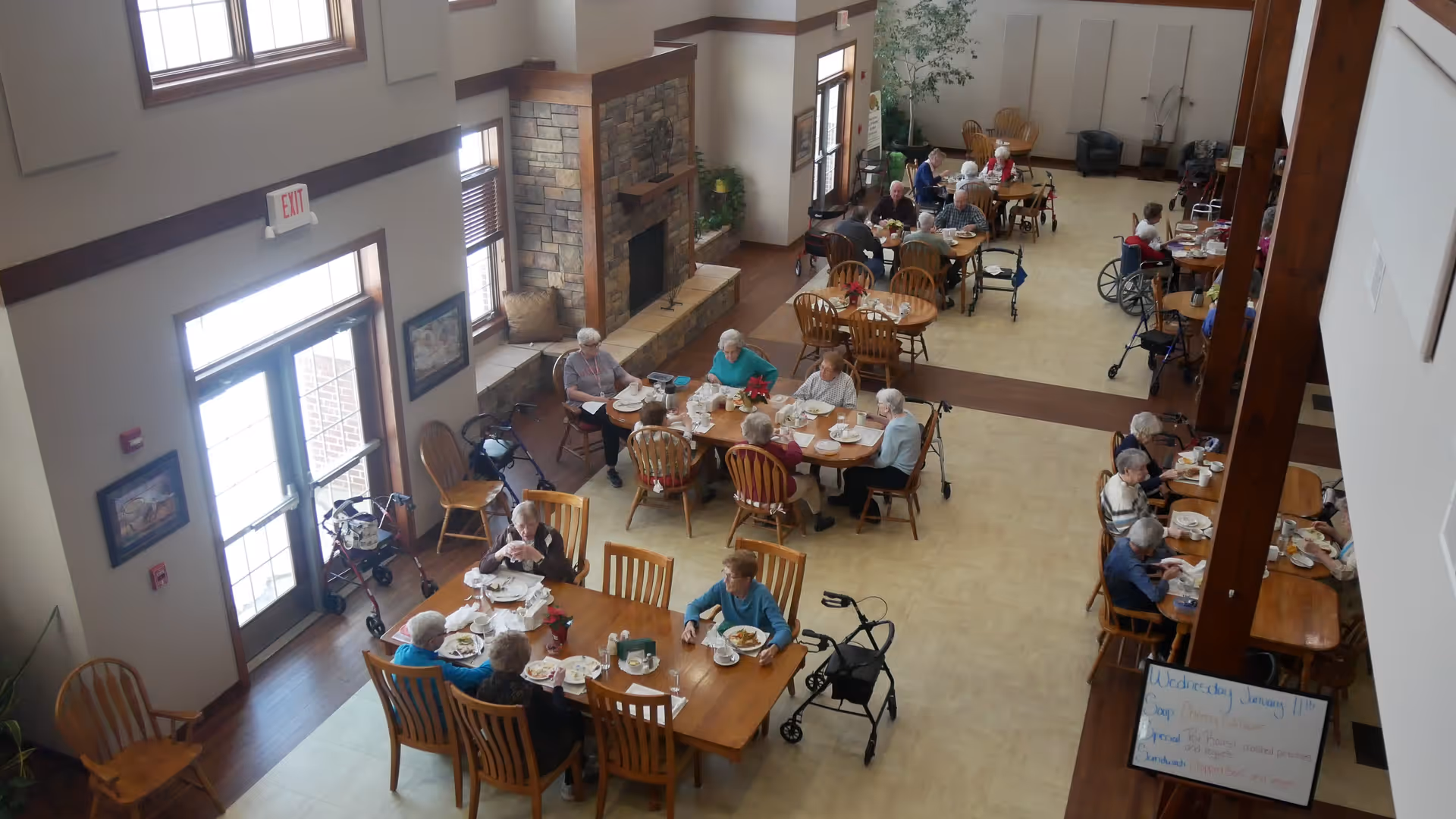 A dining room in a senior living facility with multiple elderly residents seated at wooden tables eating meals. The room has large windows, a stone fireplace, and several walkers and wheelchairs are visible near the tables. There is a whiteboard with a schedule near the entrance.