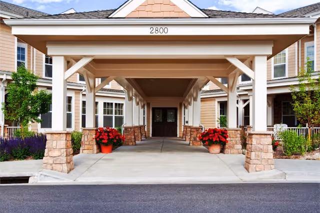 Covered entrance/porte-cochère of a senior living building with stone columns, potted red flowers, and double doors beneath the address '2800'.